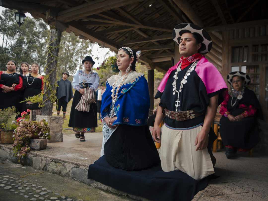 Saraguro Ceremony A Saraguro couple kneels in the foreground during a ceremony while their community observes from behind.