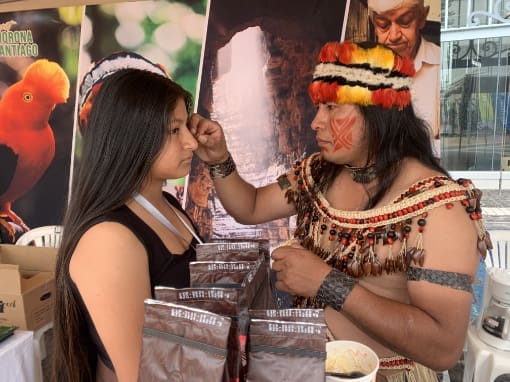 Un miembro de la nacionalidad Shuar, vestido con atuendo tradicional que incluye una banda de plumas y un collar, pinta el rostro de un visitante con achiote, en un acto de intercambio cultural y conexión ancestral.