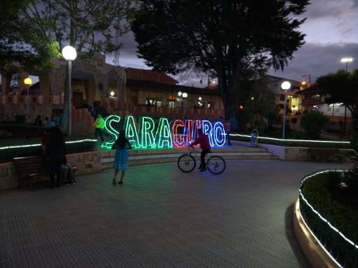 Un colorido letrero de neón con la palabra 'Saraguro' brilla intensamente en el parque frente a la iglesia al anochecer, mientras niños en bicicletas aparecen como siluetas en perfil.