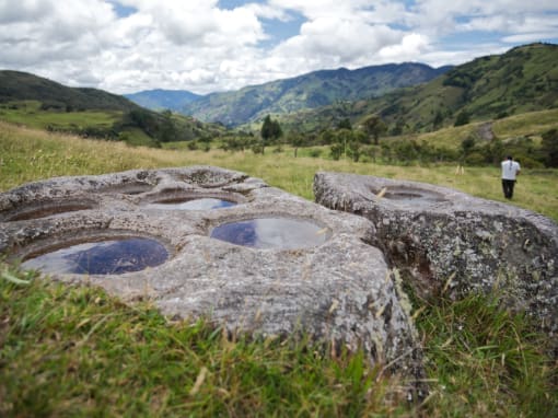 Ancestral Rocks Ancient rocks with carved basins, possibly used for grinding, lie in the middle of a pasture surrounded by mountains, as a man walks in the distance.